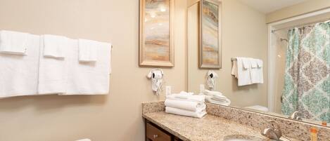 A bathroom with a granite countertop, a sink, a toilet, and a shower with a patterned curtain. There are towels hanging on the wall and a hairdryer on the counter.