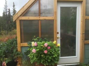A wooden greenhouse with a glass door, surrounded by lush greenery and flowering plants. - Denali Park Place - Alaskan elegance with over 350 Five Star reviews! (Denali National Park)