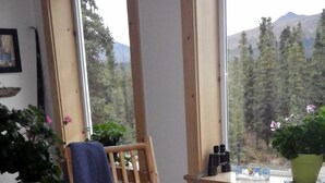 A wooden rocking chair with a blue towel, a window with a view of trees and mountains, and a small table with a potted plant.