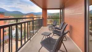 A balcony with wicker chairs and a small table, overlooking a mountainous landscape with buildings. - Premier Slopeside Apartment | Fireplace (Breckenridge)