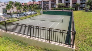 Ein Tennisplatz mit umgebender Umzäunung, neben einem Parkplatz und Wohngebäuden mit Solaranlagen.