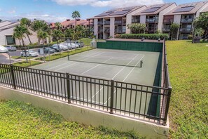 A tennis court with a surrounding fence, adjacent to a parking area and residential buildings with solar panels. - Hale Kamaole 150 | Ground-Floor Ocean View, Lanai, King Bed, Split A/C, Sleeps 4 (Kihei)