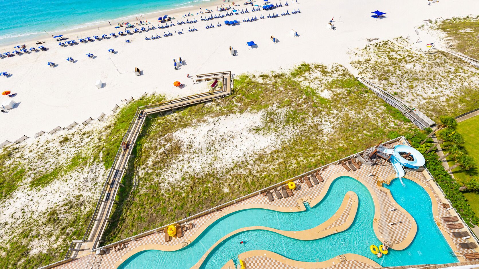 An aerial view of a beach resort with a swimming pool, beach umbrellas, and a water slide.