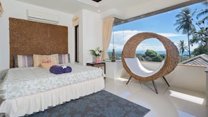 Een slaapkamer met een groot bed, een ronde rieten fauteuil en uitzicht op palmbomen en de oceaan.
