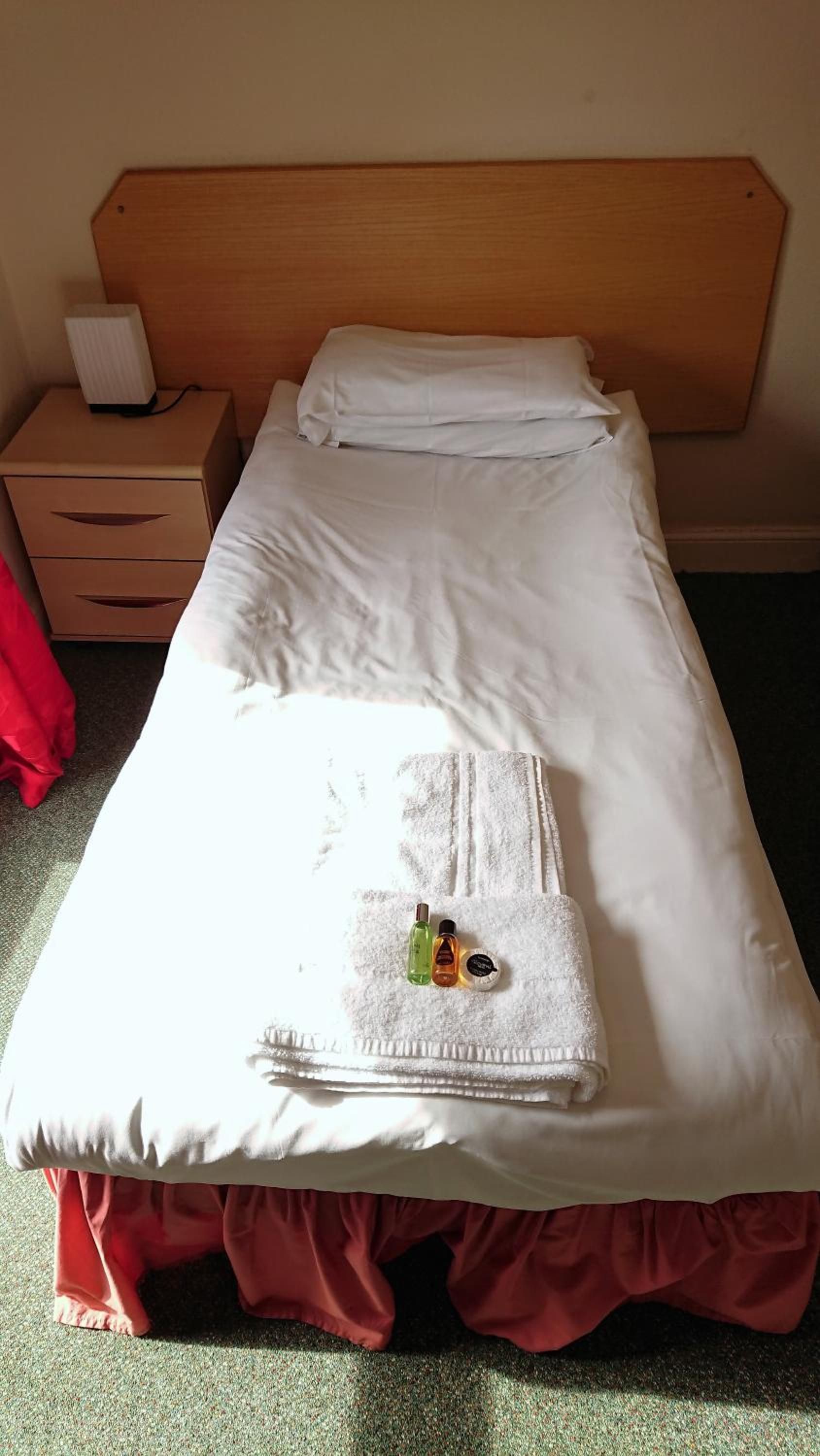 Un lit bien fait, avec du linge de lit blanc, une serviette pliée et deux flacons de produits de toilette posés sur une autre serviette blanche.