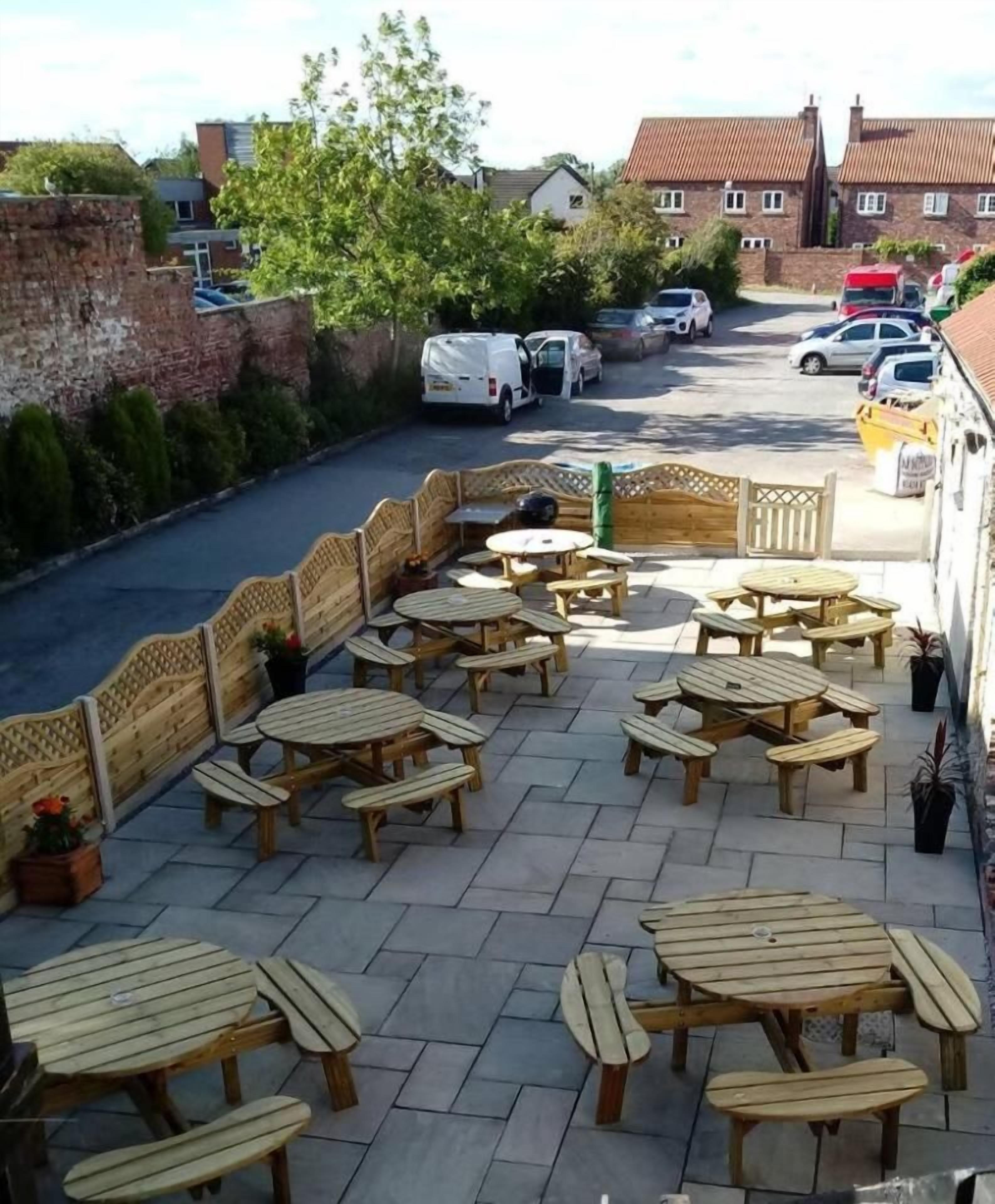 Un patio al aire libre con mesas y bancos de madera, un muro de ladrillo y vistas de la calle al fondo.