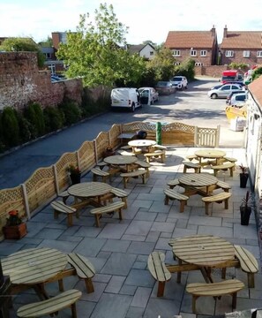 An outdoor patio area with wooden tables and benches, a brick wall, and a street view in the background. - Wellington Hotel (Goole)