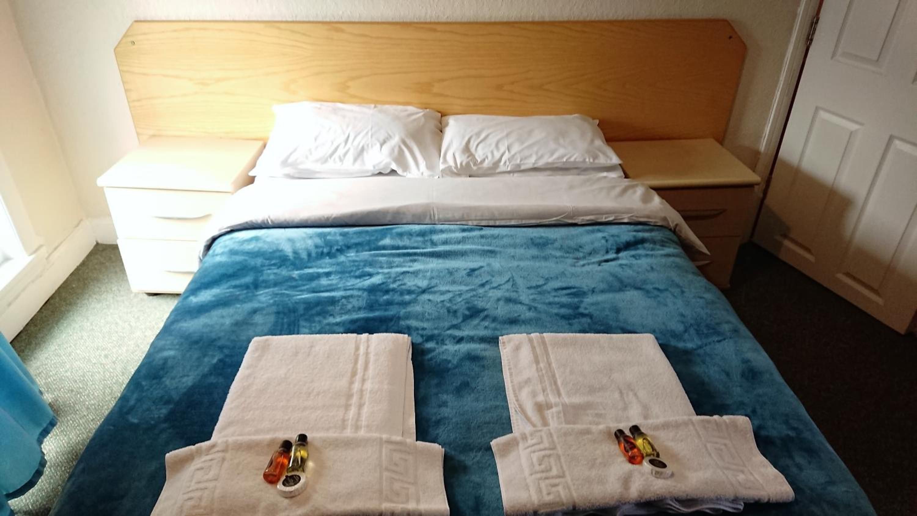 A hotel room with a wooden headboard, a blue bedspread, white pillows, and two towels with bottles on them.