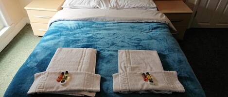 Una cama bien hecha con una manta azul y dos toallas dobladas, junto con artículos de tocador en una habitación de hotel.