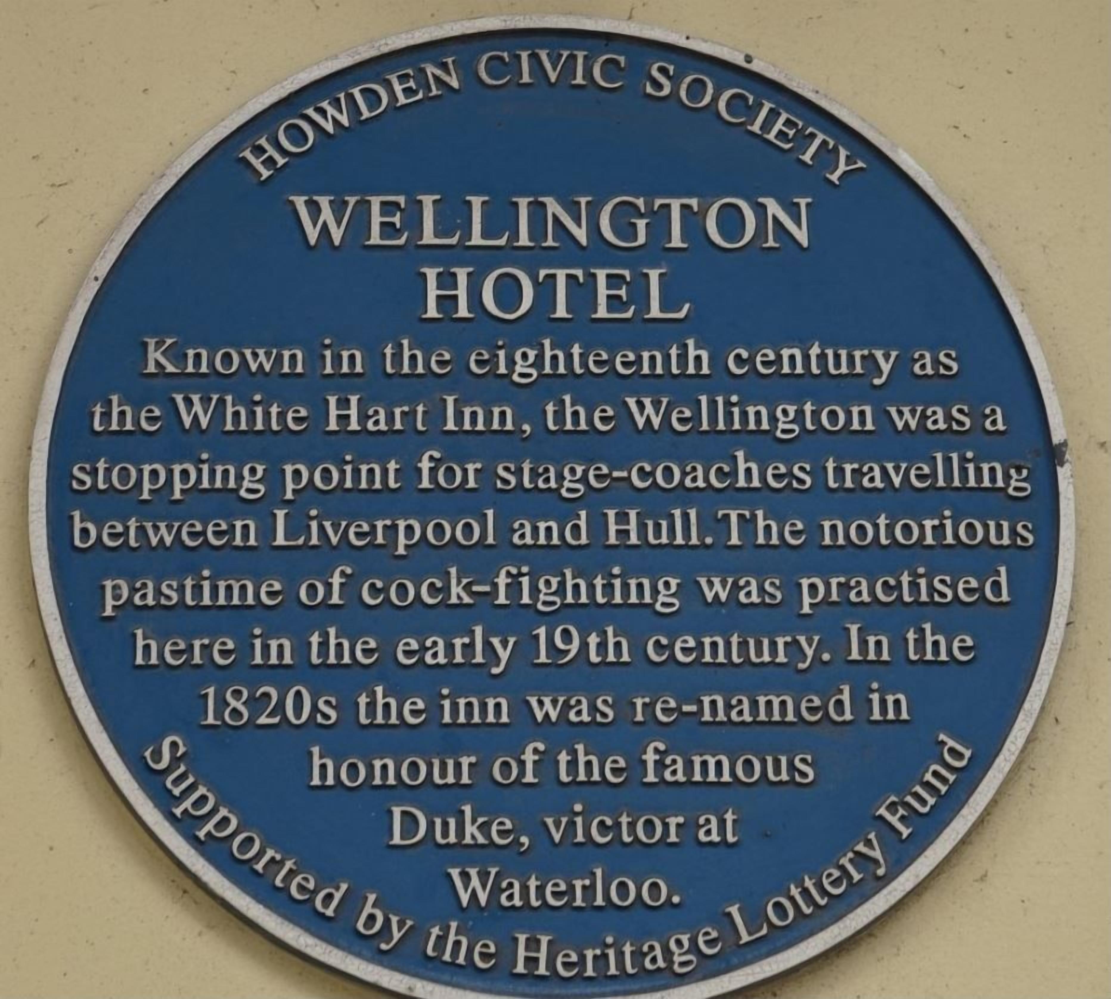 Een plaquette met de geschiedenis van Hotel Wellington, inclusief gebruik in de vroege 19e eeuw als postkoetsherberg en plaats voor hanenvechten, en de hernoeming in de jaren 1820 ter ere van de hertog van Wellington.