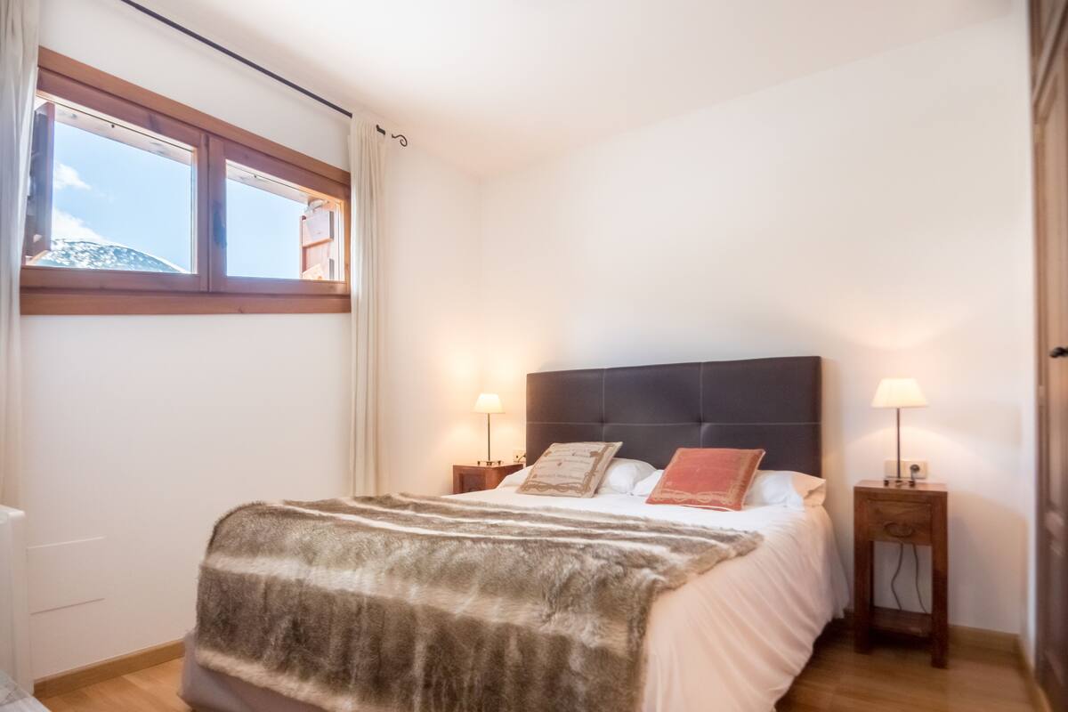 A bedroom with a large bed, two bedside tables with lamps, a window with curtains, and a view of the sky.