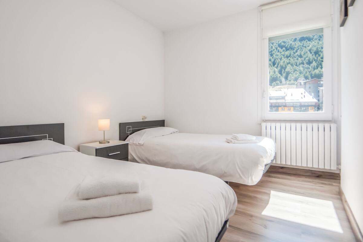 A modern hotel room with two beds, a nightstand with a lamp, and a window with a view of a forest and buildings.