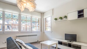 Una sala de estar moderna con sofá, mesa, televisión y ventanas amplias que ofrecen vistas a edificios y vegetación.