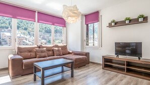 Una sala de estar moderna con un sofá de cuero marrón, una mesa de centro de madera y un televisor de pantalla plana sobre un mueble de madera. Amplias ventanas con persianas rosadas ofrecen vistas a un paisaje montañoso.