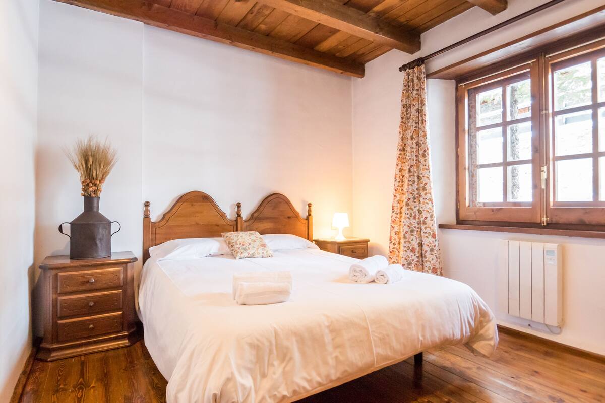 Ein Schlafzimmer mit einem hölzernen Bett, einem Nachttisch mit Lampe, einer Kommode und einem Fenster mit Vorhängen.