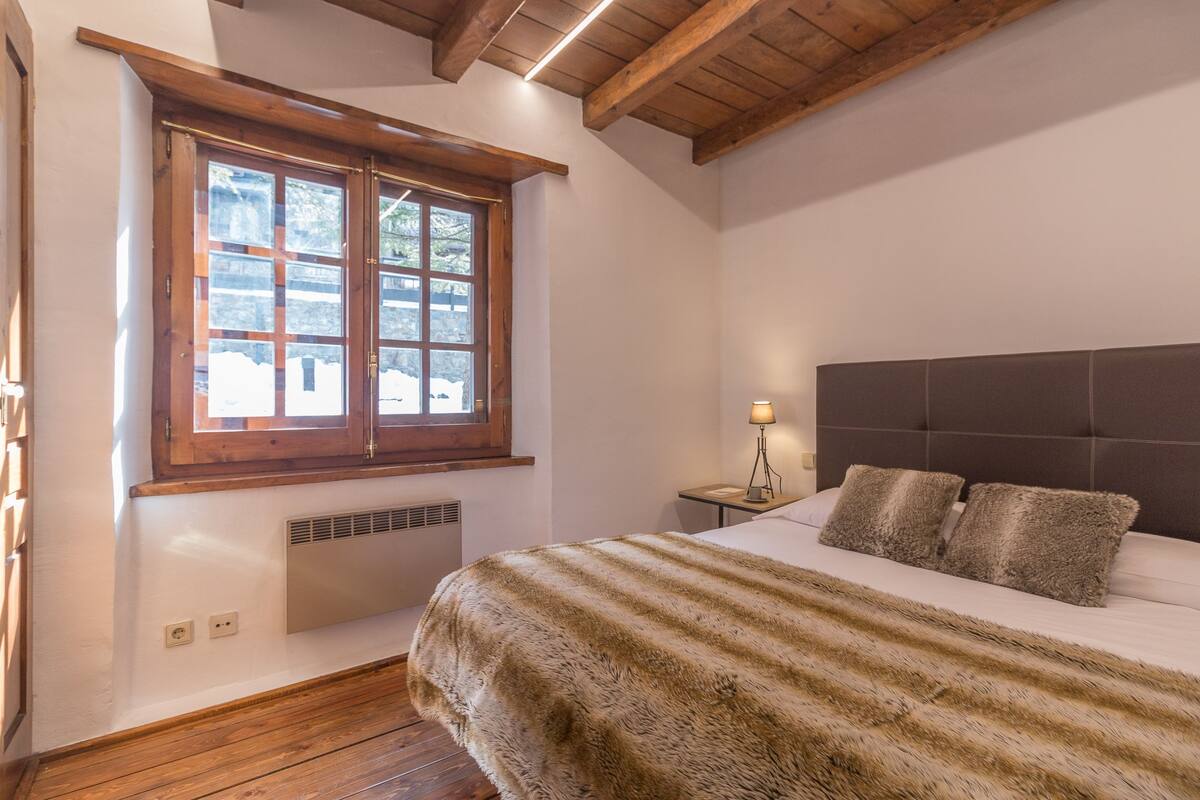 Ein Schlafzimmer mit einem großen Bett, Holzbalken an der Decke, einem Fenster mit Blick und einem Nachttisch mit Lampe.