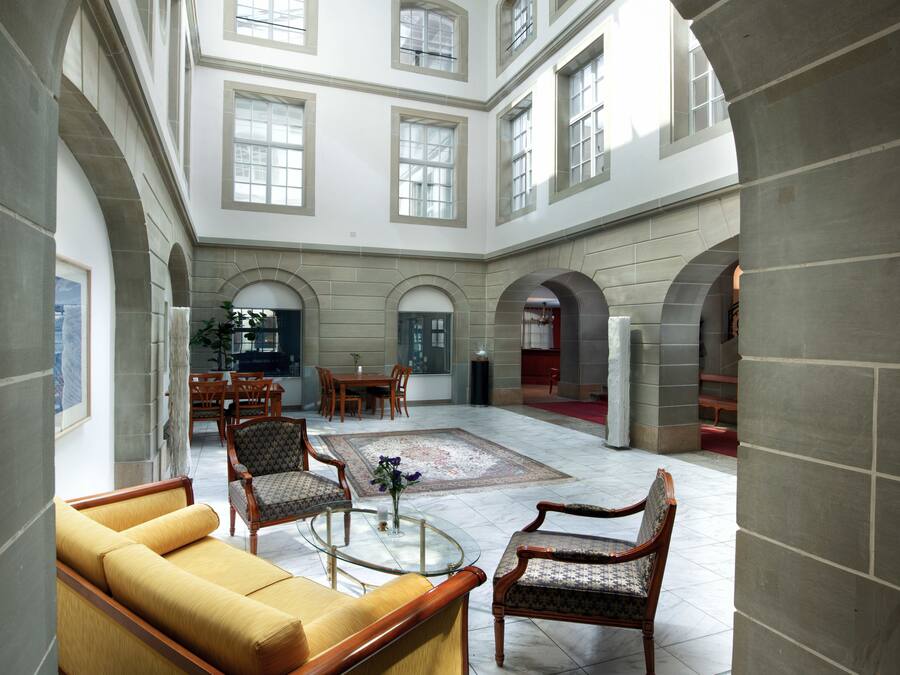 Eine geräumige Lobby mit hoher Decke, großen Fenstern und einer zentralen Sitzecke mit Sofas und Sesseln.
