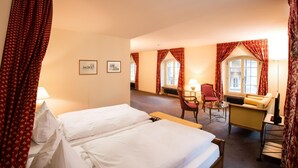 A hotel room with a bed, a chair, a sofa, and a table. There are windows with curtains, a radiator, and framed pictures on the wall. - Hotel Stadthaus (Burgdorf)