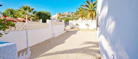 A narrow gravel pathway leading to a white-walled building with a decorative fence and palm trees in the background.