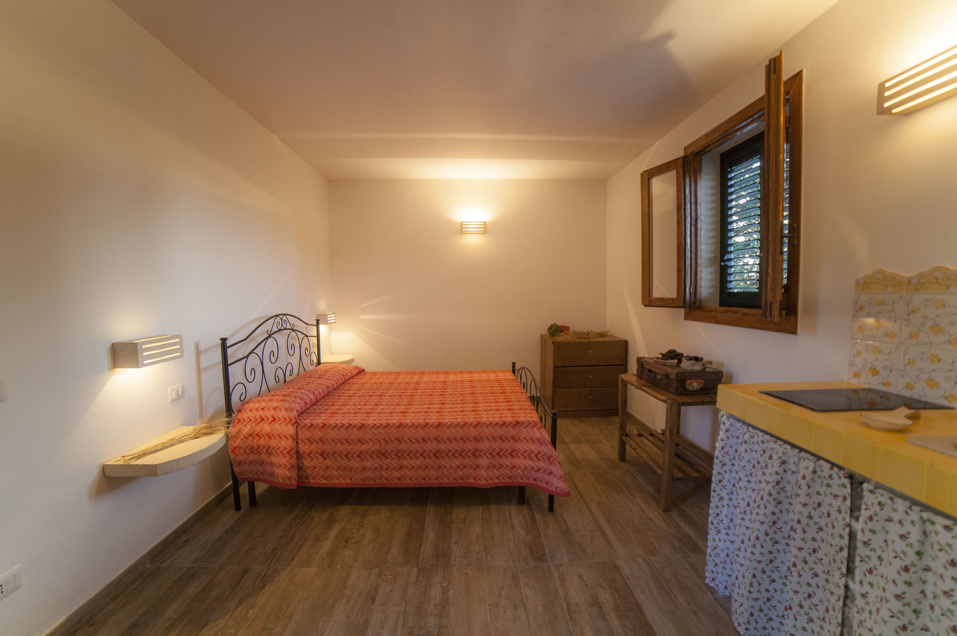 A small, well-lit bedroom with a bed, bedside table, and a window with blinds.
