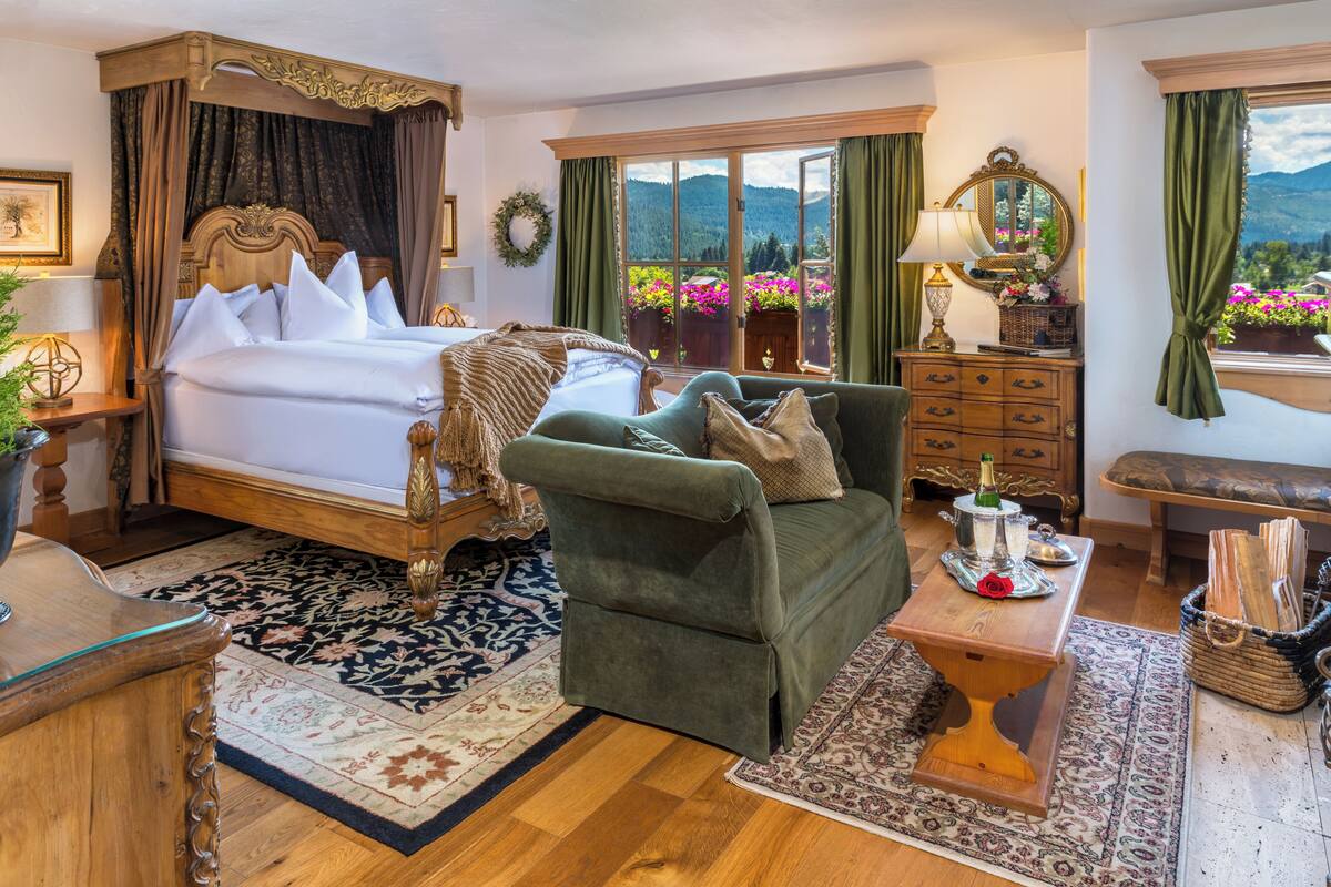 Une chambre à coucher avec un lit à baldaquin, un fauteuil vert, une commode en bois et une vue sur les montagnes par de grandes fenêtres.