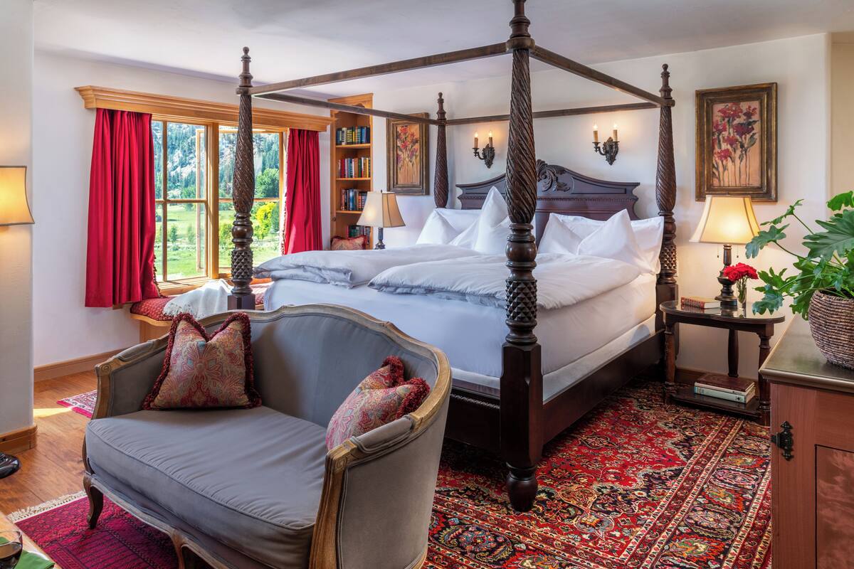 Un lit à baldaquin avec un couvre-lit à motifs rouges et blancs, une chaise en bois avec un coussin, un tapis à motifs rouges et une bibliothèque contenant des livres et une lampe.