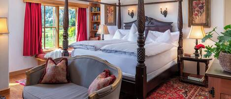 A four-poster bed with a red and white patterned throw, a wooden chair with a cushion, a red patterned rug, and a bookshelf with books and a lamp.