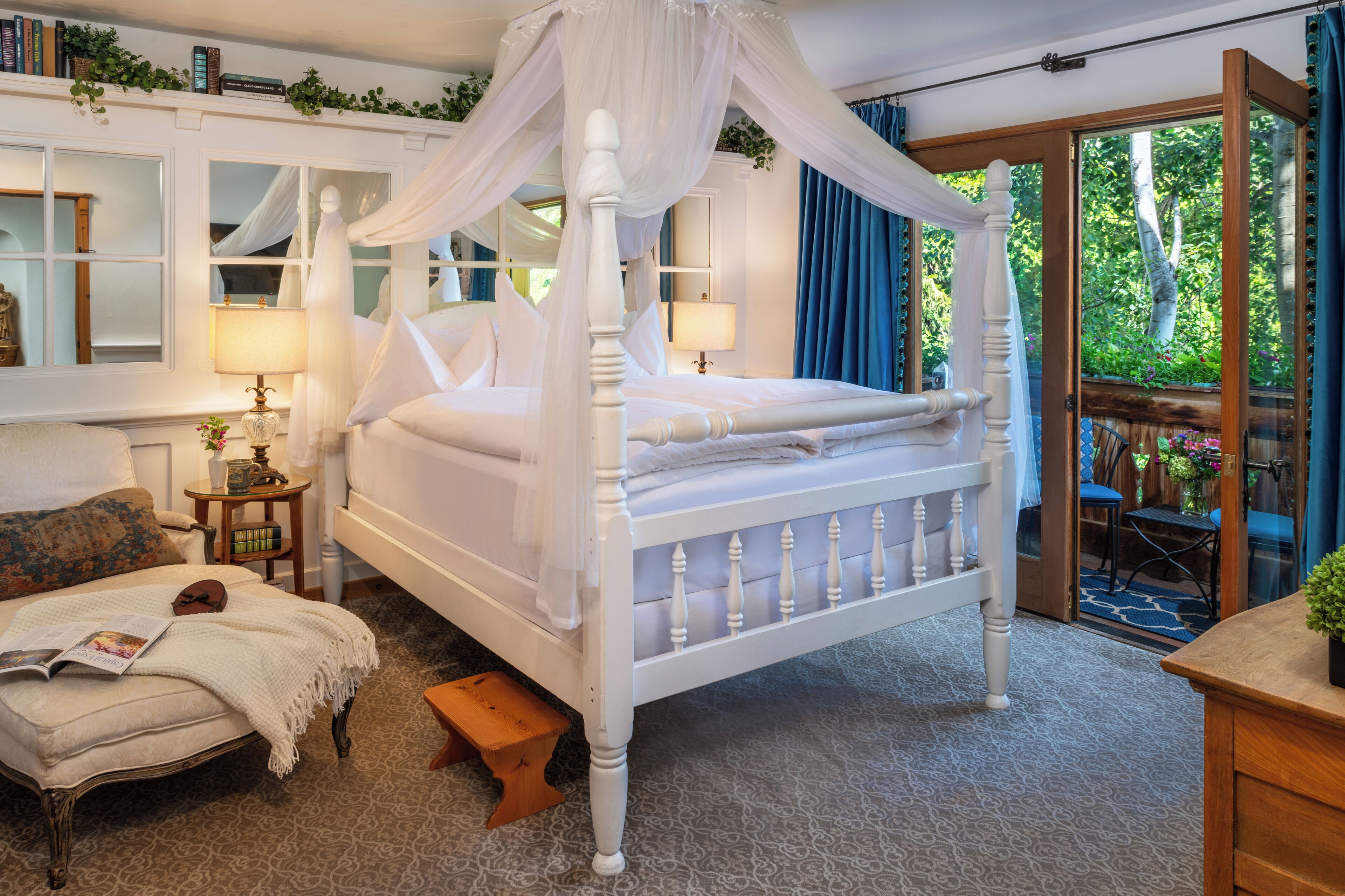 A four-poster canopy bed with a white finish, a wooden bench, a small table with a lamp, and a view of the outdoors through a glass door.