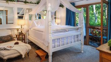 A four-poster canopy bed with a white finish, a wooden bench, a small table with a lamp, and a view of the outdoors through a glass door.