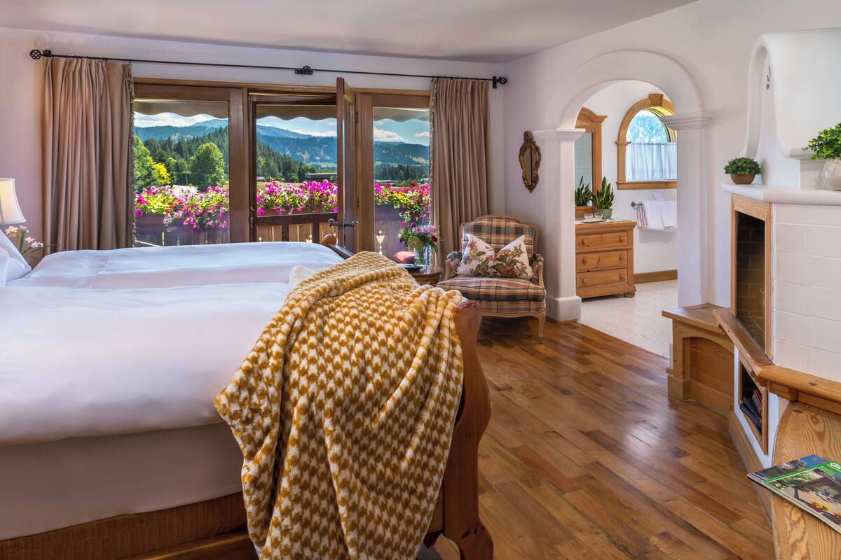 Une chambre avec un grand lit, un plancher en bois, une vue sur les montagnes et les fleurs par la fenêtre, une chaise, une commode et une cheminée.