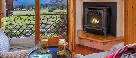 A cozy room with a fireplace, a balcony with a view of mountains, and two chairs with a table in the foreground.