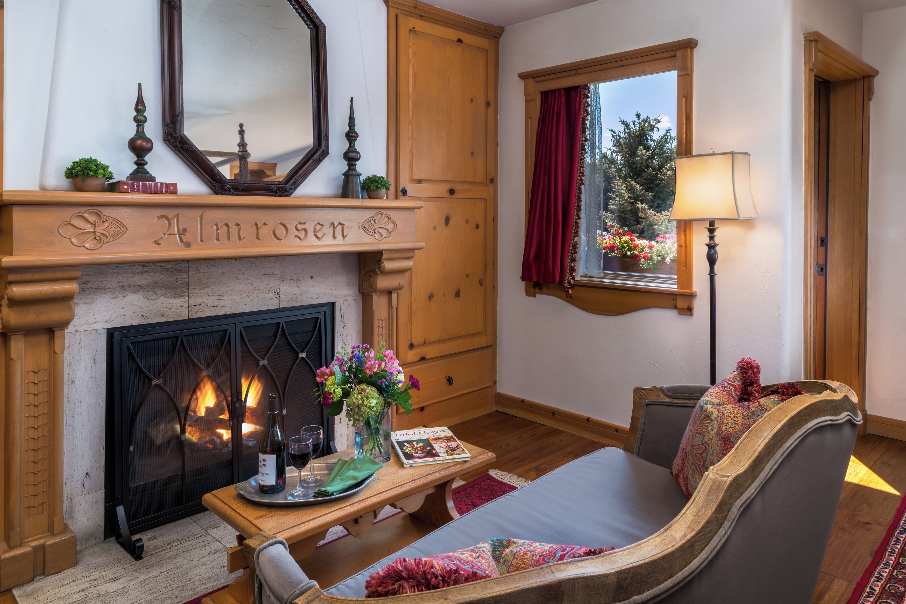 A cozy living room with a fireplace, a sofa, a table with a book and a bottle, and a window with a view of trees.
