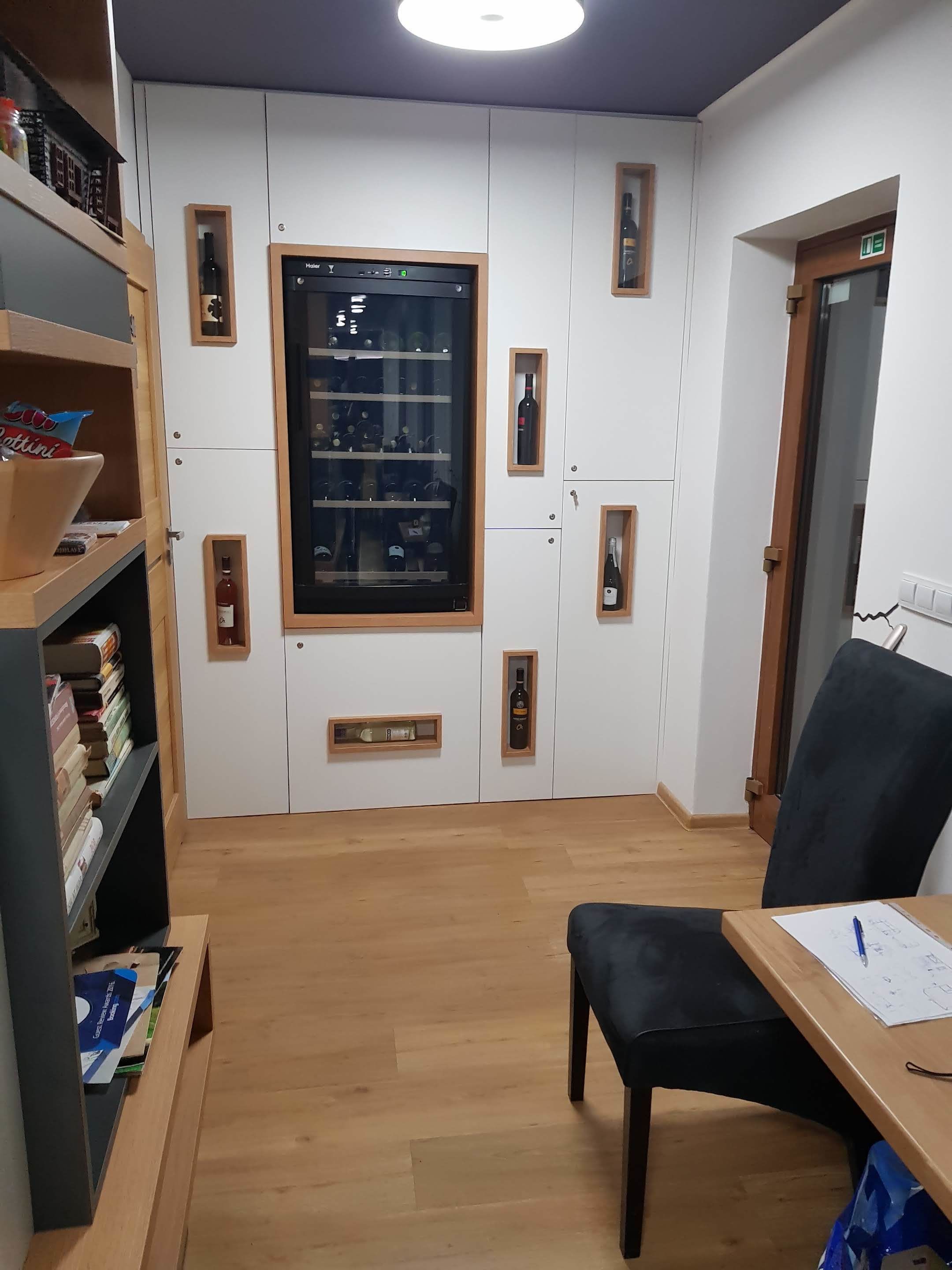 A room with a wooden floor, a desk with papers, a chair, and a shelf with books. There is a large window with a wine rack and a door with an emergency exit sign.