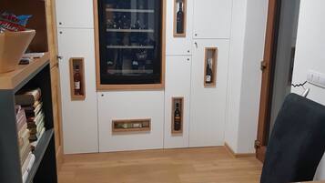A room with a wooden floor, a desk with papers, a chair, and a shelf with books. There is a large window with a wine rack and a door with an emergency exit sign.
