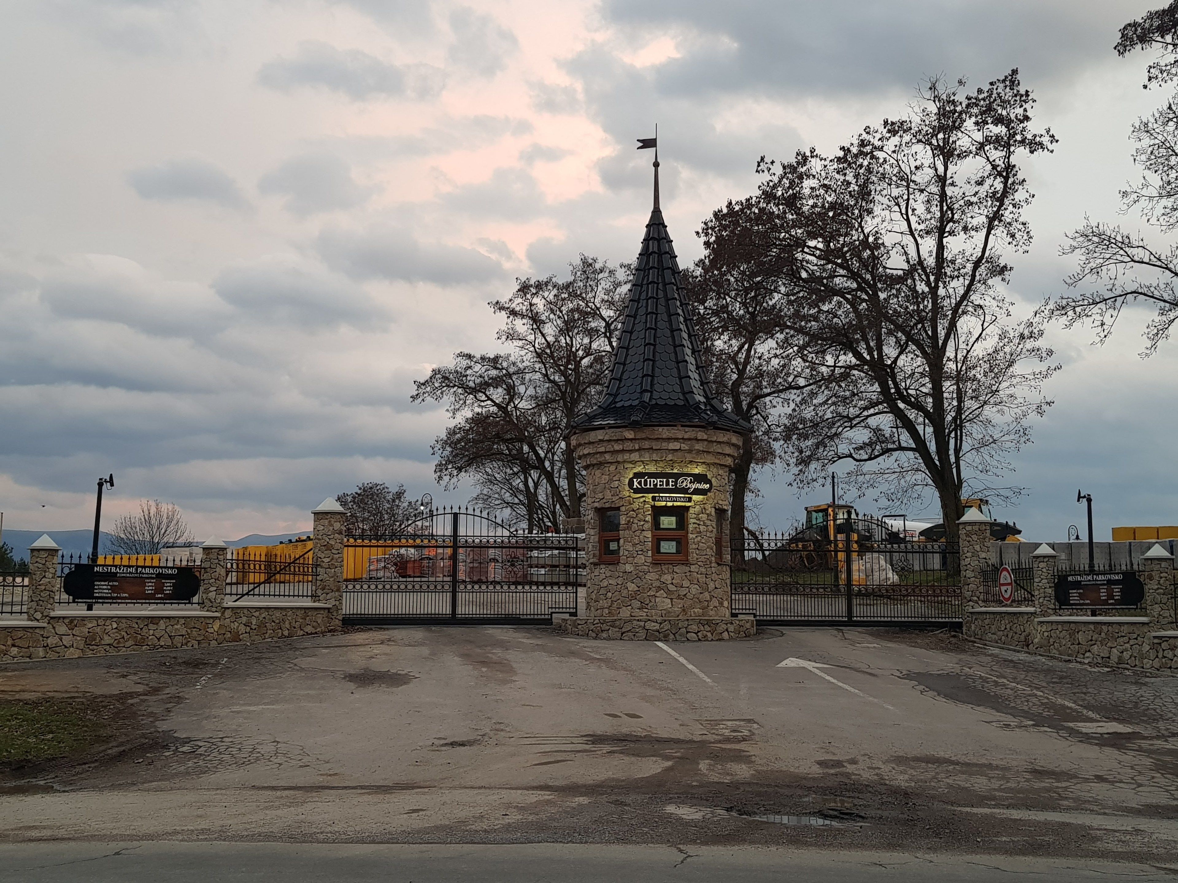 A stone building with a pointed roof and a flagpole, surrounded by a metal fence and trees.