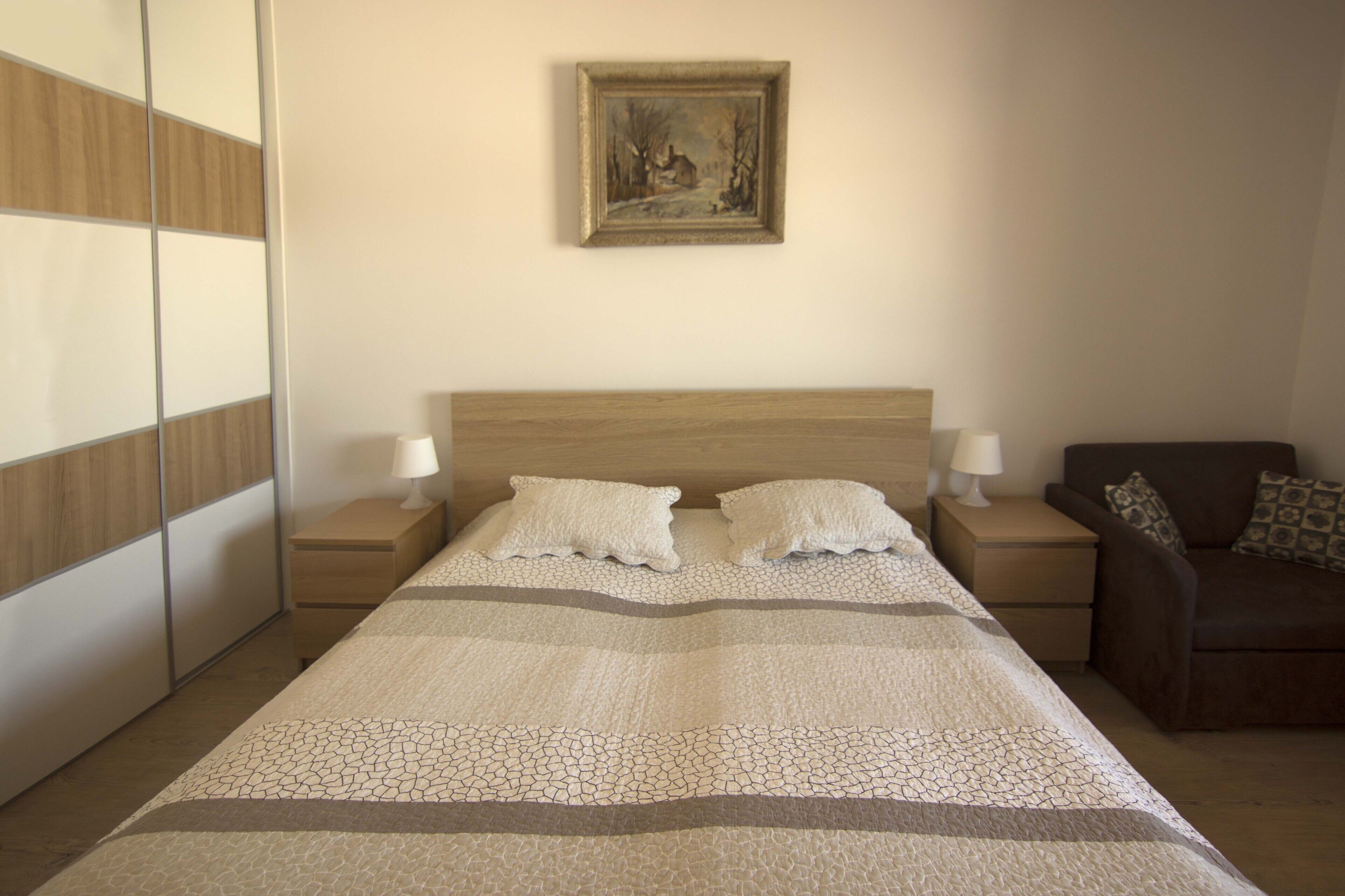 A bedroom with a large bed, two bedside tables with lamps, a brown sofa, and a painting on the wall.