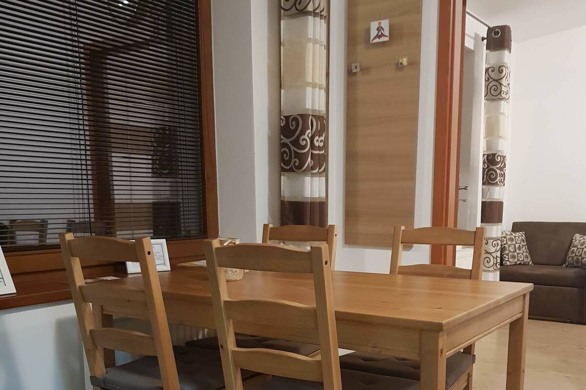 En matplats med ett träbord och stolar, ett fönster med persienner och en dörr med en dekorativ gardin.
