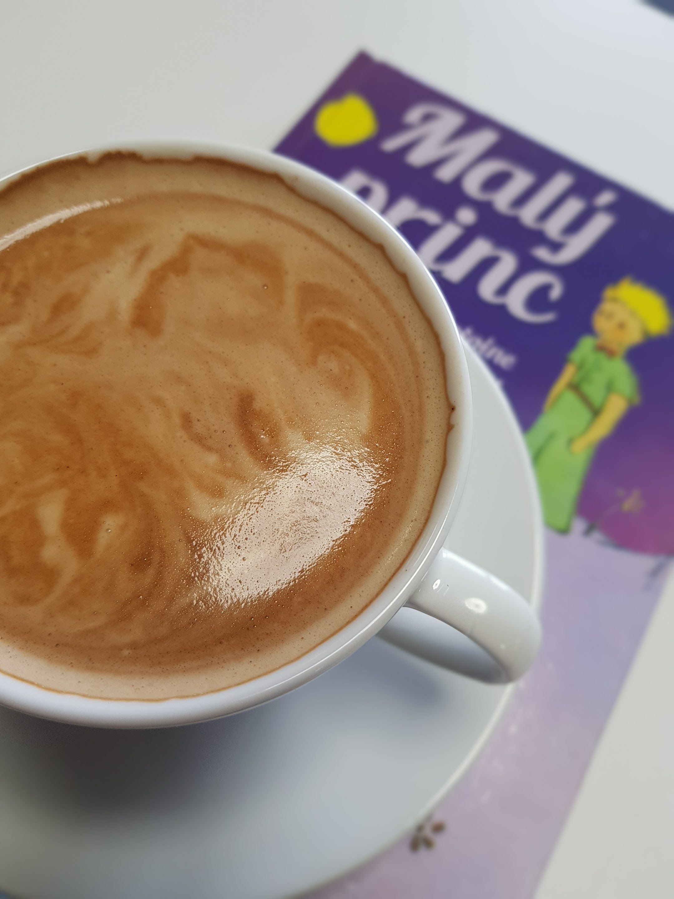 Een kop koffie met latte-art op een schoteltje, geplaatst op een tafel met een paars en geel brochure.