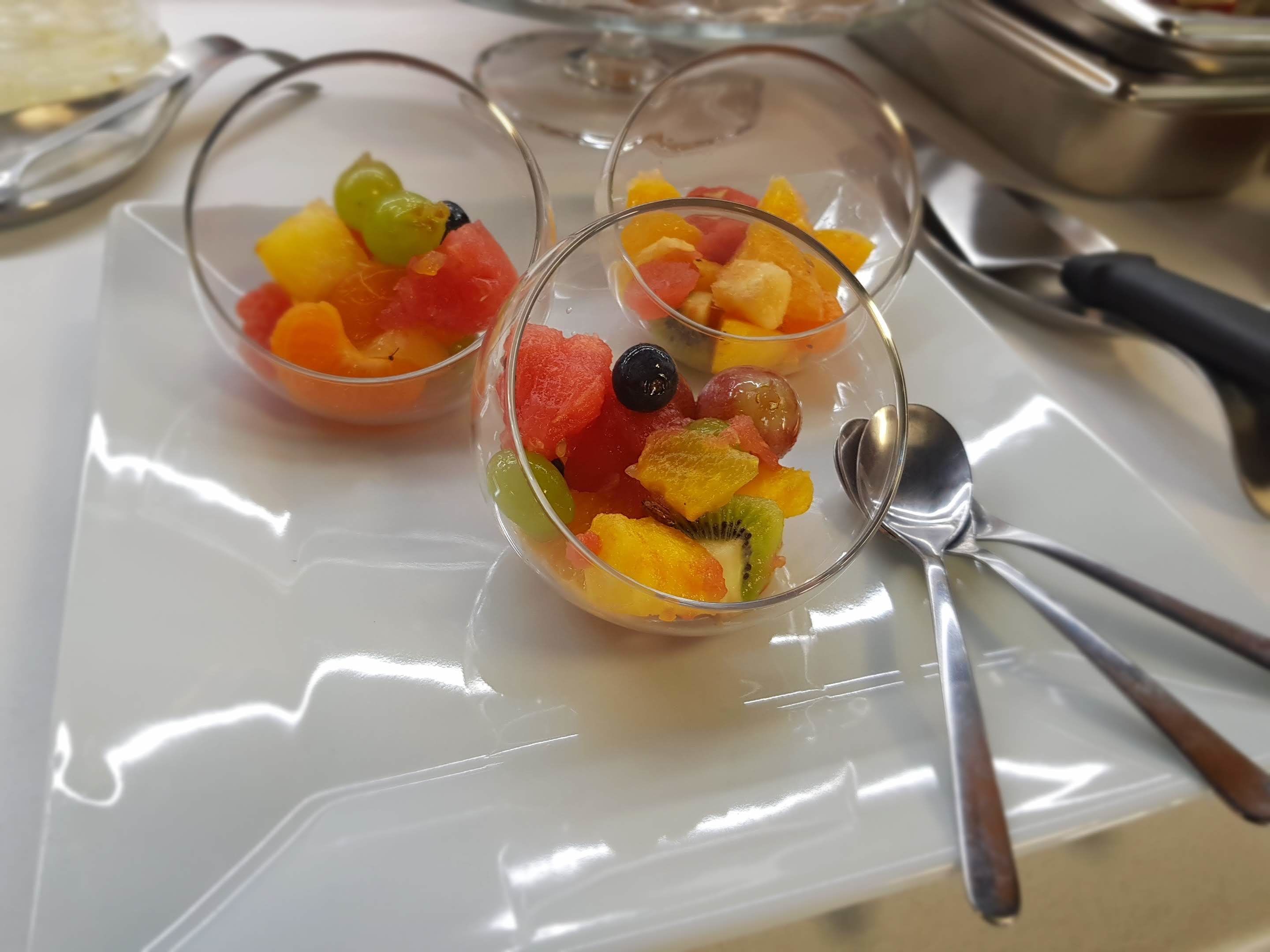 Tres recipientes de vidrio transparente repletos de frutas variadas sobre una bandeja reflectante.