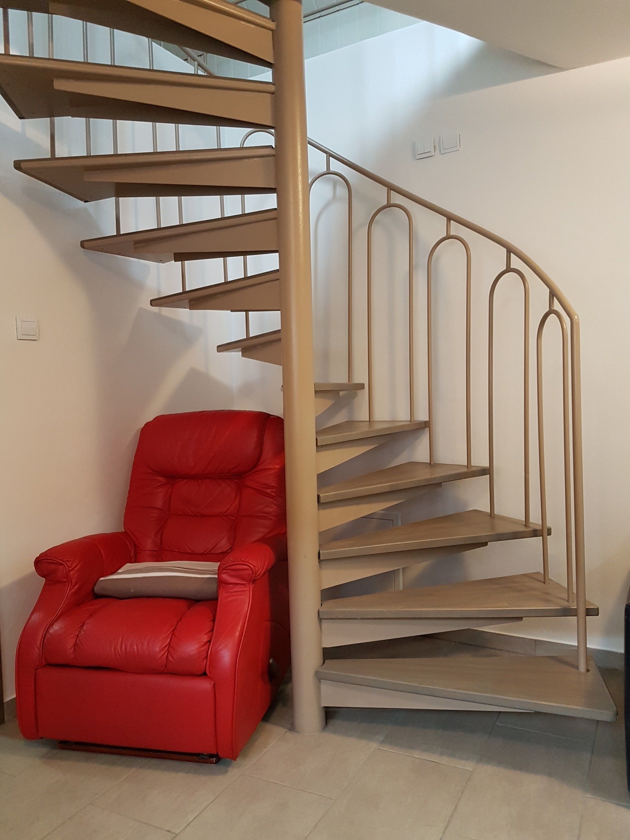 Un escalier en colimaçon moderne, avec une finition beige, un fauteuil en cuir rouge doté d’un coussin et un plancher de couleur claire.