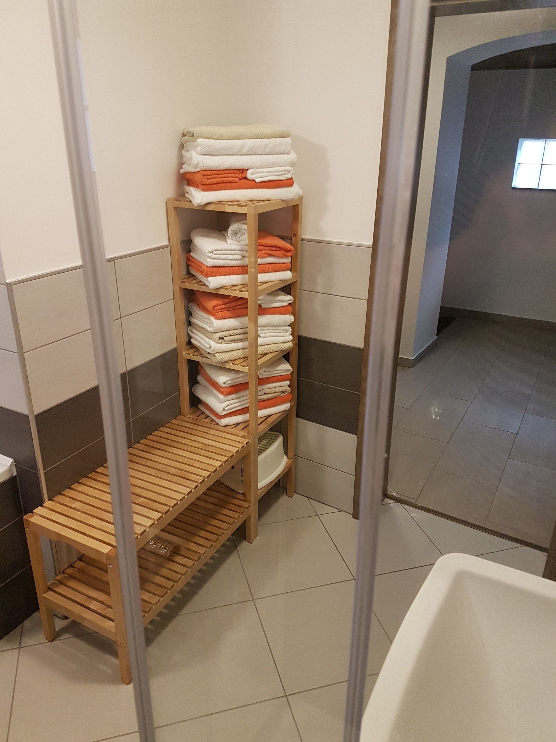 A bathroom with a wooden shelf holding folded towels, a wooden bench, and a white bathtub.