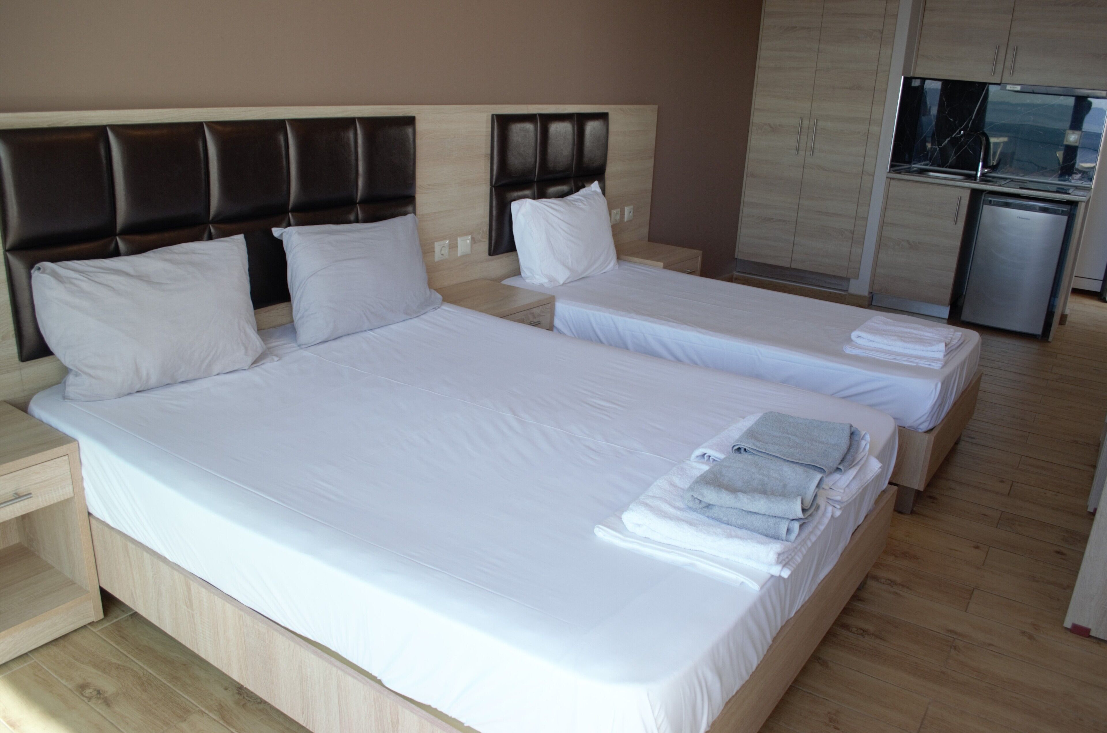 A hotel room with two beds, a wooden headboard, a small kitchen area, and a wooden floor.