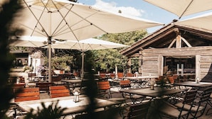 An outdoor patio with wooden tables and benches, shaded by large umbrellas, and a rustic wooden building in the background.