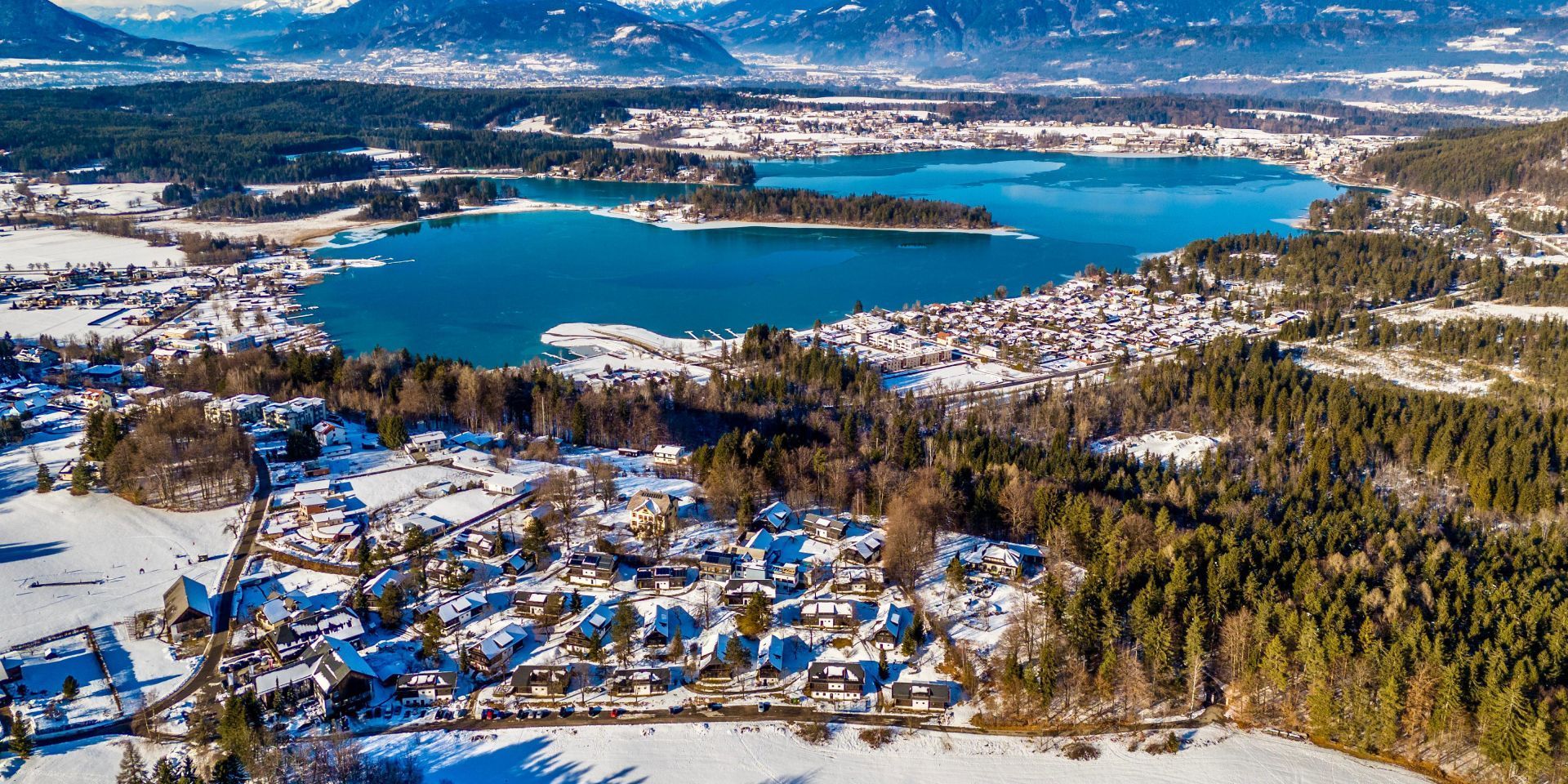 An aerial view of a winter town with a lake, surrounded by snow-covered houses and dense forests.
