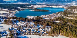 Vista aérea de un pueblo invernal con un lago, rodeado de casas cubiertas de nieve y densos bosques.