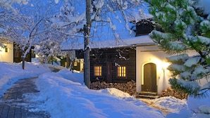 A snow-covered pathway leading to a wooden cabin with a lit entrance and windows.