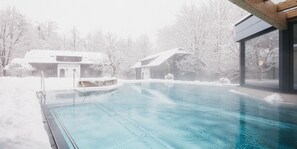 Una piscina con cerramiento de vidrio, rodeada de árboles y edificios cubiertos de nieve.