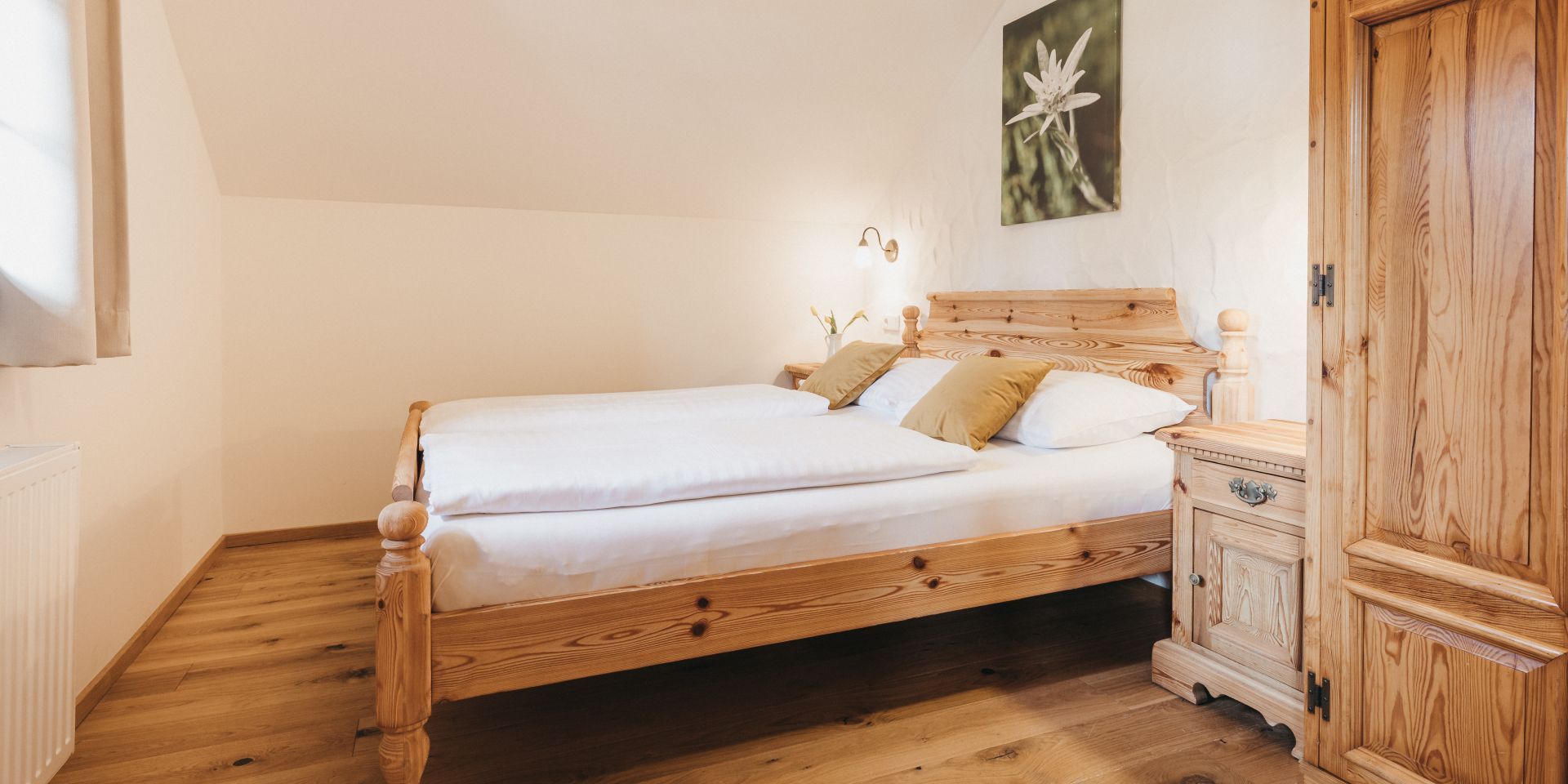 A wooden bed with white bedding, a nightstand with a lamp, and a wooden wardrobe.