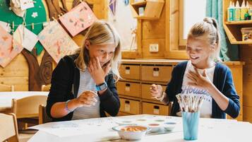 Two people at a table with art supplies, wooden cabinets, and a bulletin board in the background.