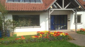 Una casa con tetto in tegole rosse, un'ampia finestra e una porta blu. Davanti, un giardino fiorito e colorato.